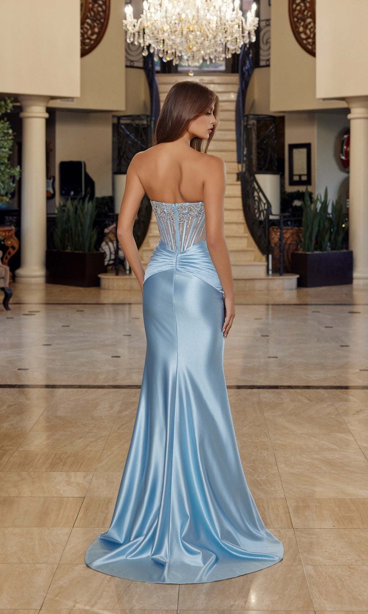 A woman wearing the Nox Anabel R1820 long prom dress, featuring a strapless fitted corset bodice and a light blue satin skirt, stands with her back to the camera in an elegant, marble-floored interior.